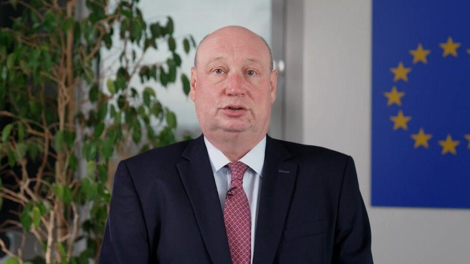 Henrik Hololei head and shoulders in front of green plant and EU flag