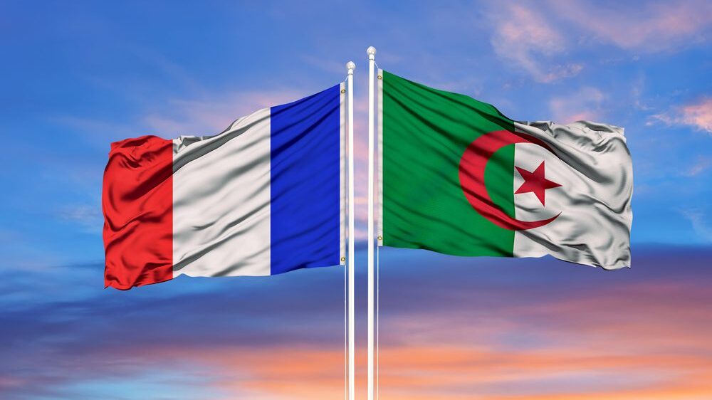 Algerian and French flag on two flag poles, wind blowing them in different directions against sunset background