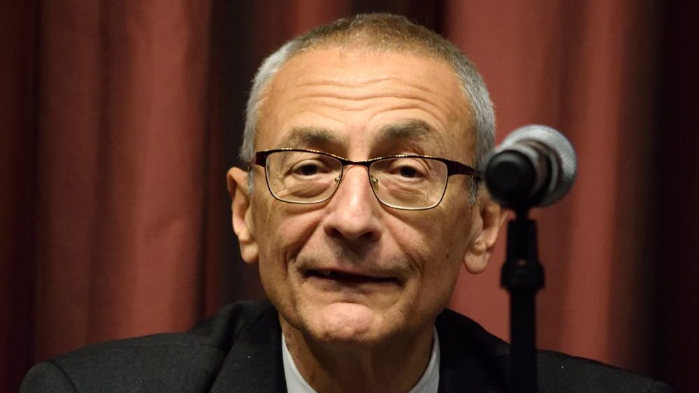 John Podesta headshot by microphone against dark red backdrop