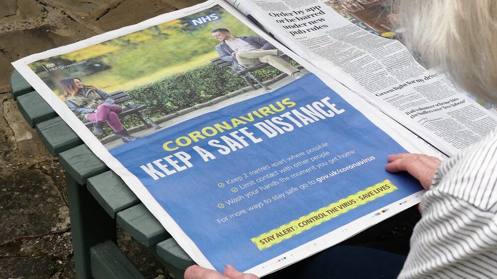 Newspaper page with government keep the distance covid ad held by gray-haired older woman