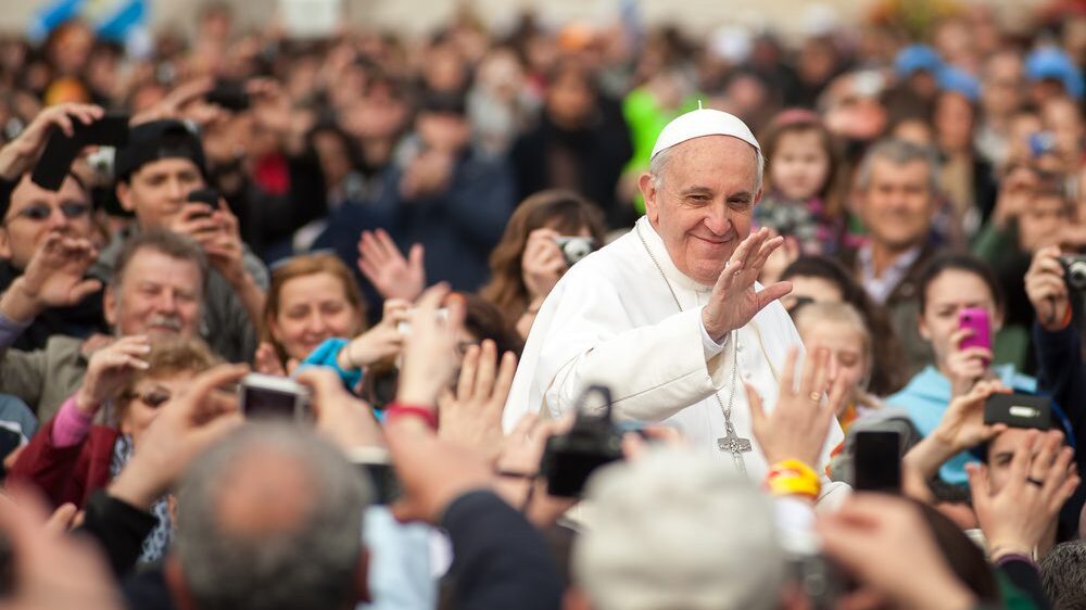 Pope Francis elevated head and shoulders above crowd smiling and waving.