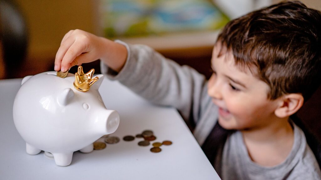 Young boy smiling while putting coins into piggybank illustrating that money has a cultural value beyond functioning as payment.