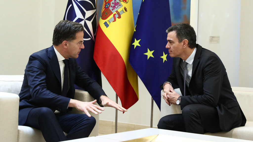 NATO chief Mark Rutte and Spanish PM Pedro Sánchez seated in armchairs facing each other talking against backdrop of NATO, Spain, and EU flags