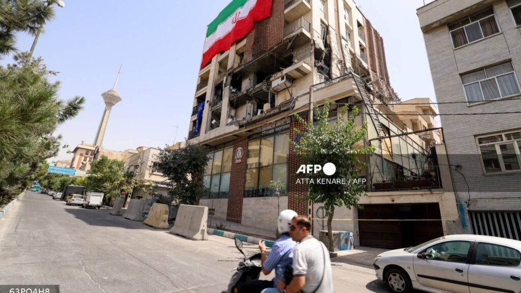 Two men ride a scooter past a residential building, hit in an Israeli strike, covered with a large Iranian flag in Tehran on June 25, 2025.