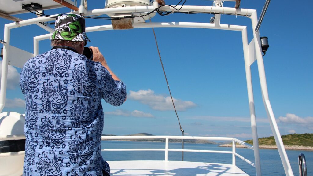 Man in Hawaiian shirt seen from behind photographing beach and ocean from aboard boat