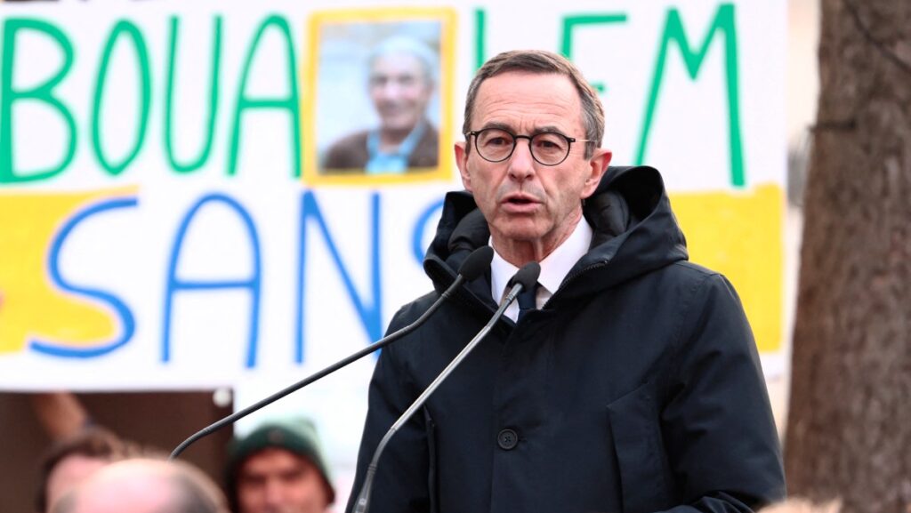 Bruno Retailleau speaking into microphone in front of a poster with the name and photo of Boualem Sansal