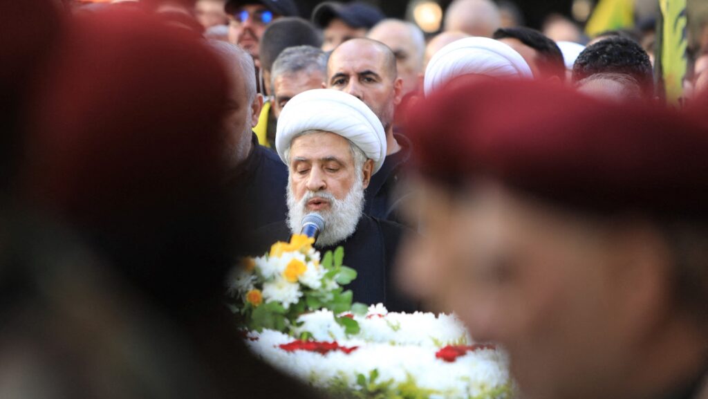 Face of Naim Qassem seen above flowers on coffin between unidentifiable faces