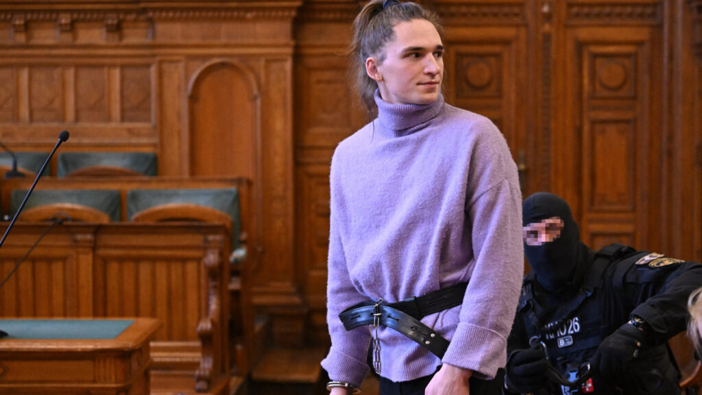 Man with ponytail and lavender turtleneck detained by belt and handcuffs in court room