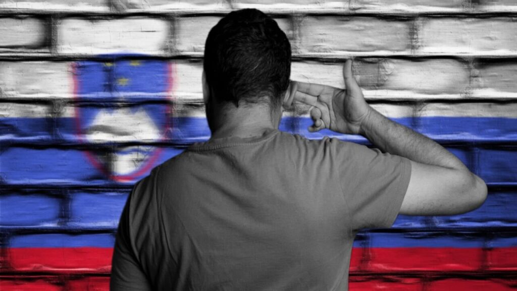 Man seen from behind, holding two fingers to side of his head as if holding a gun, in front of brick wall with Slovenian flag projected onto it.