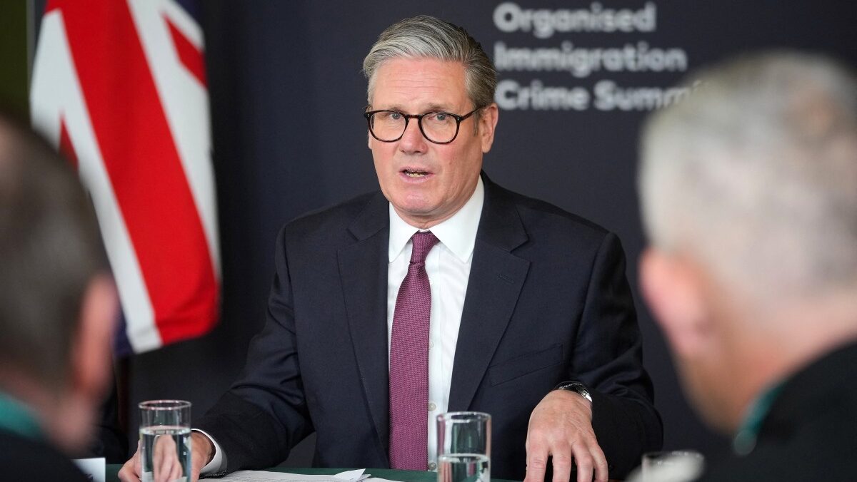 Starmer seated at table talking with UK flag and text on wall saying "Organized Immigration Crime Summit" behind him.