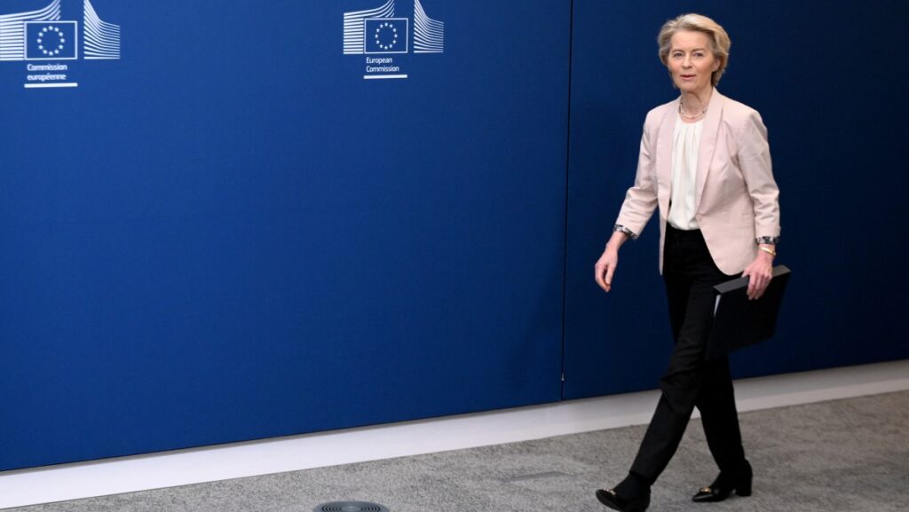 von der Leyen walking with light smile on her face against backdrop of blue wall with EU Commission logo