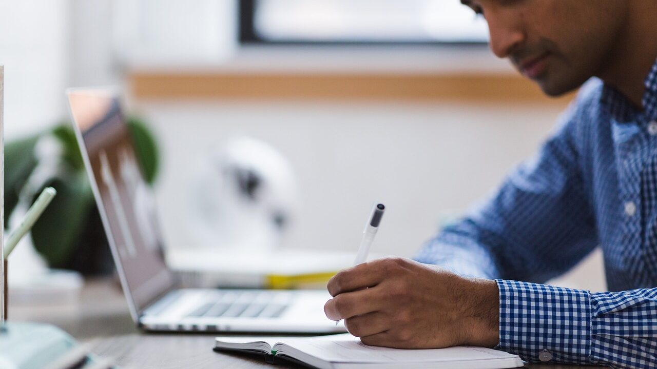 dark-skinned man in front of laptop taking notes with pen and paper notebook