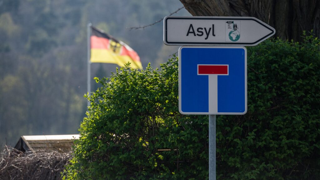 End of the road sign with arrow pointing right saying Asylum in German, German flag in the back