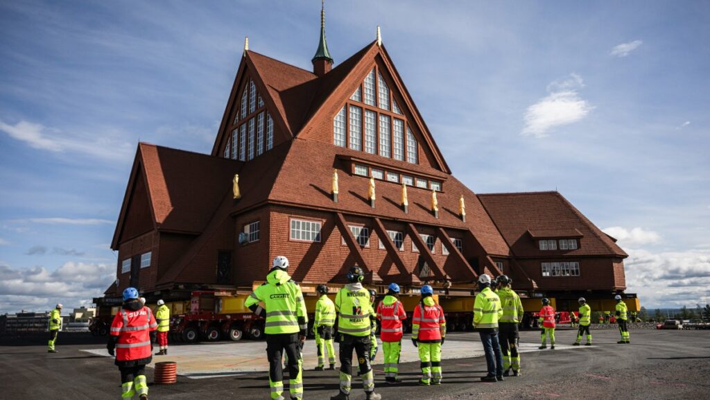 Large wooden church sitting on many transportation trucks with staff in hi-vis vests watching