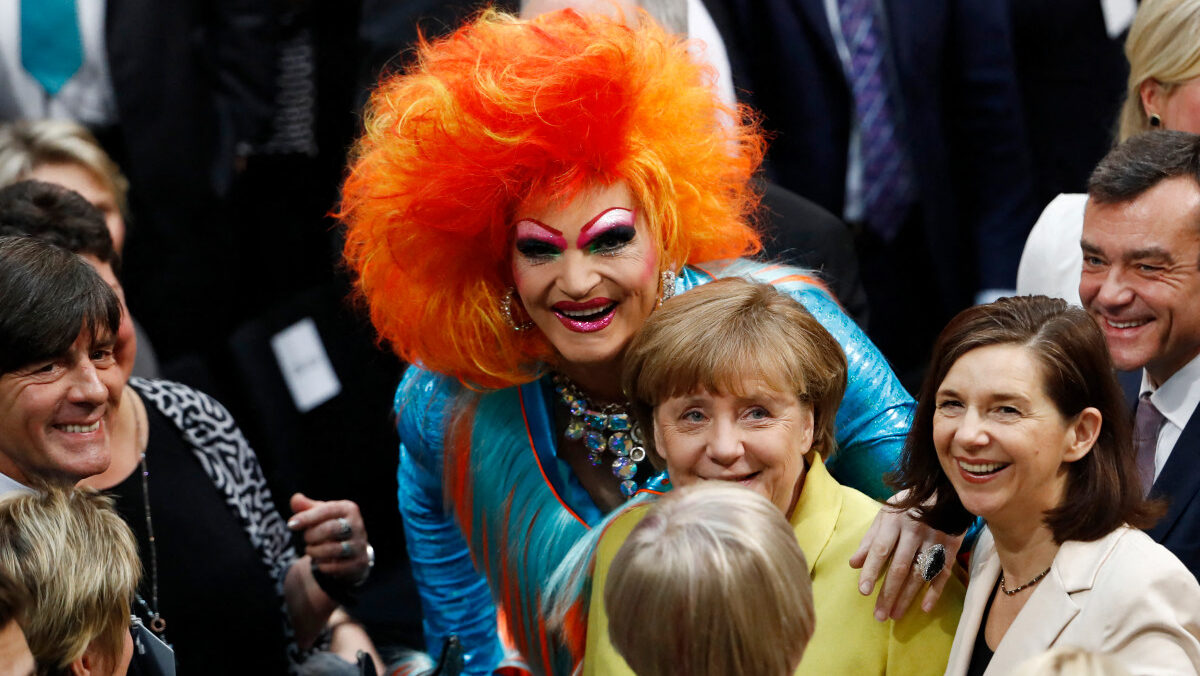 Drag queen with large orange hair with his hand on a smiling Merkel's shoulder