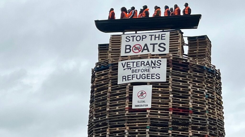 Models depicting migrants wearing life jackets in a small boat alongside two banners reading 'Stop the boats' and 'Veterans before refugees' are displayed on top a bonfire in Moygashel, Northern Ireland, on July 9, 2025.