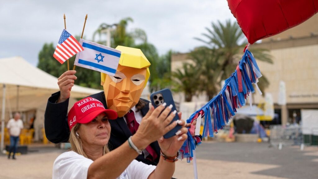 A person wearing a mask depicting U.S. President Donald Trump holds US and Israeli flags after the hostage deal declared by Trump, at Hostage Square in Tel Aviv on October 9, 2025.