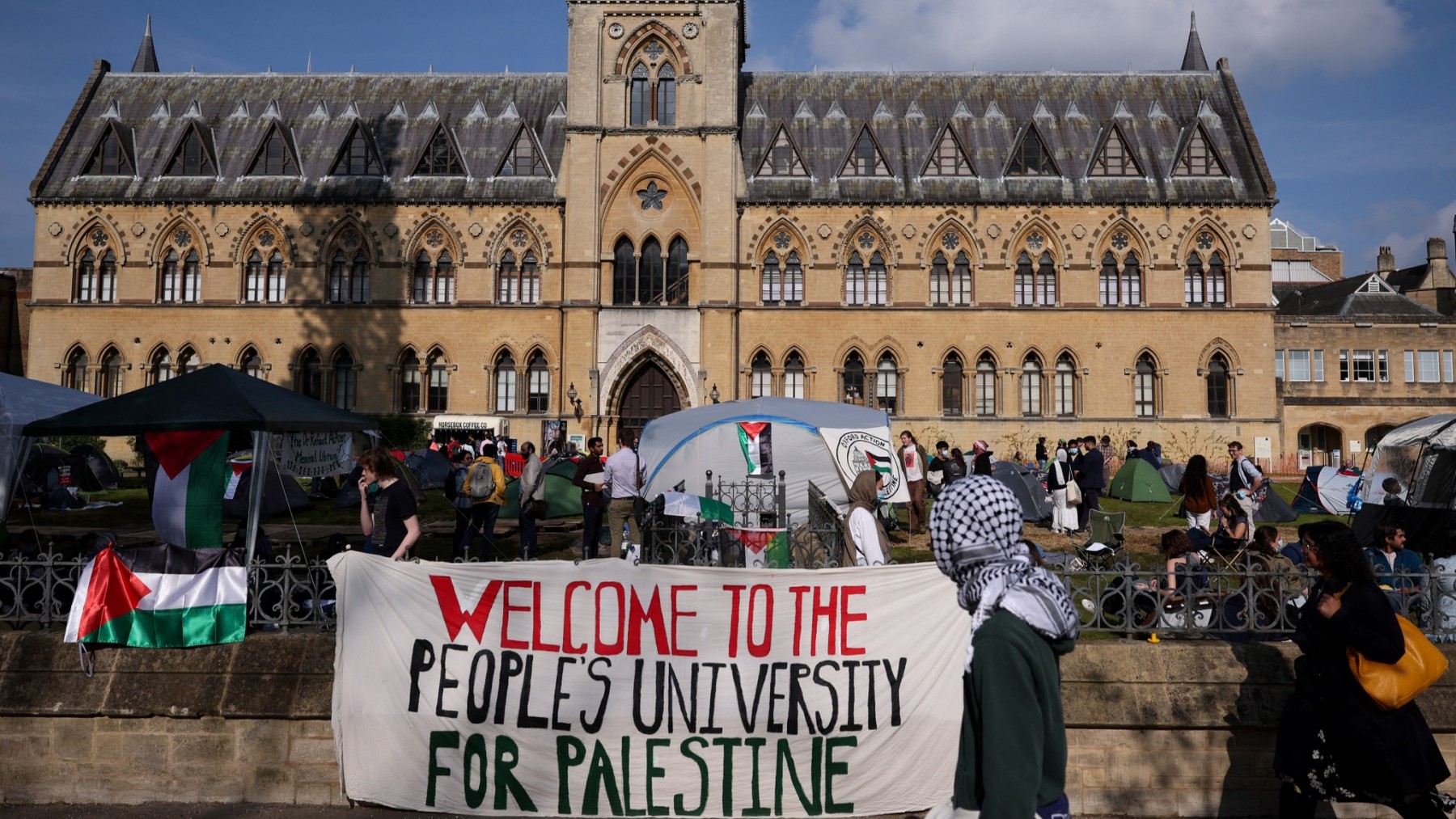 A passerby with the head covered with a keffiyeh walks past a banner reading "Welcome to the people's university for Palestine" at Oxford University, in Oxford on May 7, 2024, as a pro-Palestinian camp has been set up on the campus.