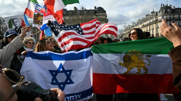 Anti-Iranian regime protesters wave a giant pre-revolutiona Iranian flag (R), an Israeli flag (L) and a U.S. flag (rear C) as they gather during a march in support of the U.S.-Israeli attack on Iran and the killing of Iran’s supreme leader, in Paris on March 1, 2026.
