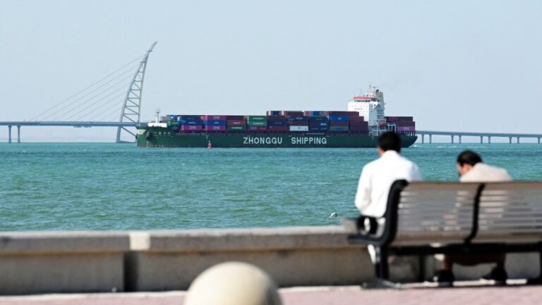 People sit on a bench on the Sheikh Jaber al-Ahmad al-Sabah Causeway as a Zhonggu Shipping container ship sails past on its way towards the port of Shuwaikh in Kuwait City on February 28, 2026.