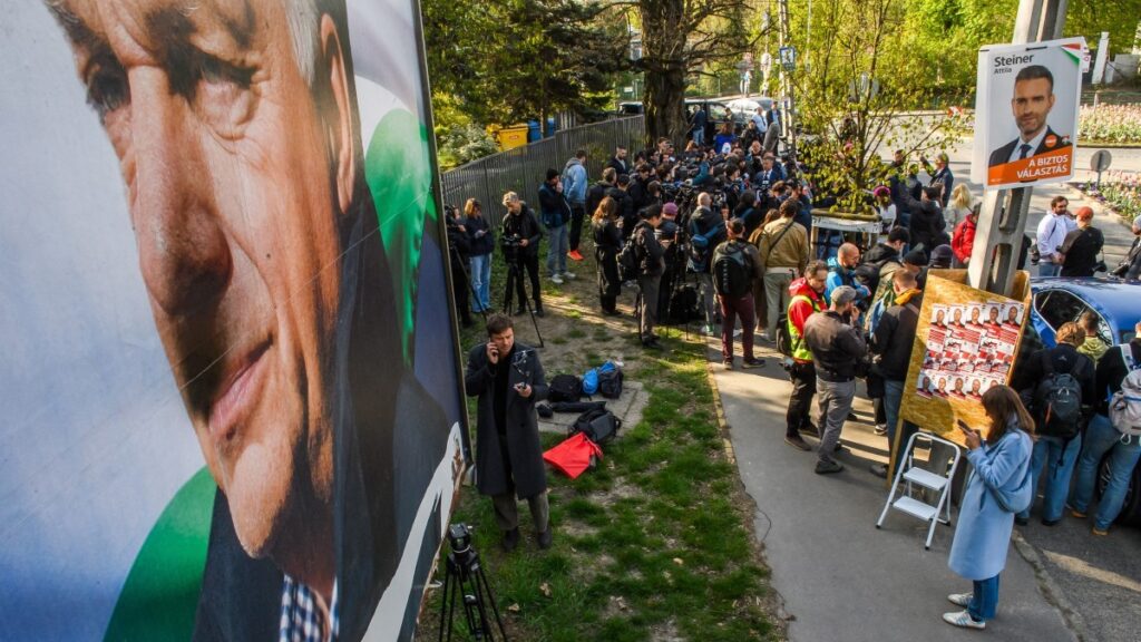 Tisza Party leader Péter Magyar (background, C) talks with journalists after casting his ballot at a polling station in Budapest, with an election poster of Hungary’s Prime Minister Viktor Orbán in the foreground during the parliamentary elections in Hungary, on April 12, 2026.