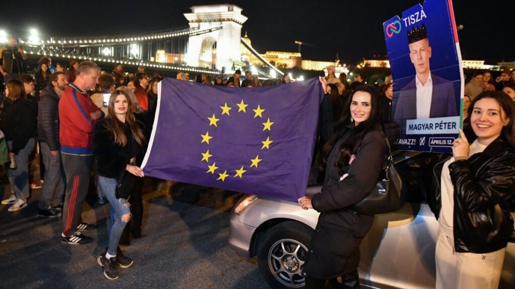 Participants display an election campaign poster of Hungary’s prime minister-elect, Péter Magyar and a flag of the European Union during a gathering to celebrate the victory of the Tisza Party near the Széhenyi Chain Bridge in Budapest late on April 13, 2026, one day after Hungary’s parliamentary elections.