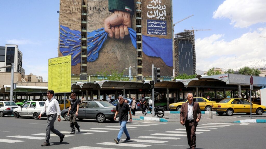 People cross a street near a billboard on the facade of a building depicting the Strait of Hormuz with a caption in Persian reading "Forever in Iran’s Hand", at Vanak Square in Tehran on May 6, 2026.