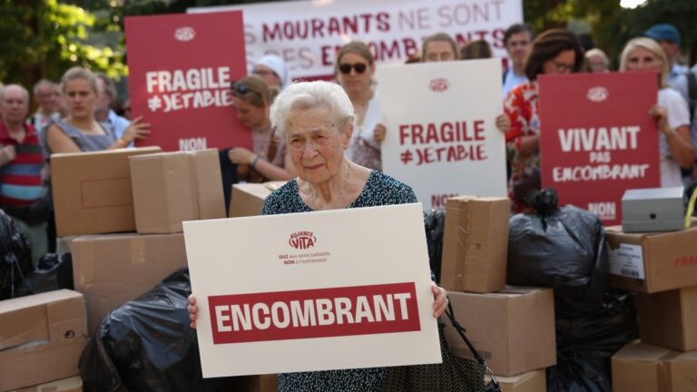 A protester sits with a placard that reads “Burden” during a rally organised by French pro-life organisation Alliance Vita against the new French draft law on euthanasia and assisted suicide at Invalides, in Paris on June 22, 2026.