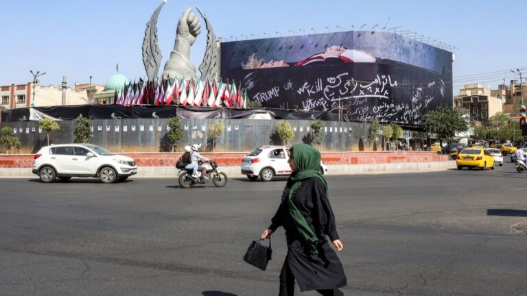 A woman crosses Tehran’s Enghelab Square on July 15, 2026, past a giant anti-U.S. billboard featuring U.S. president Donald Trump in a coffin with text in Persian reading “We Kill Trump.”
