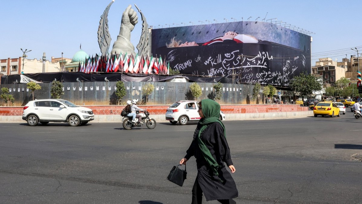 A woman crosses Tehran’s Enghelab Square on July 15, 2026, past a giant anti-U.S. billboard featuring U.S. president Donald Trump in a coffin with text in Persian reading “We Kill Trump.”